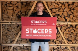 Rode doos met het logo van STOCERS wordt vastgehouden door eigenaar Cars Bijlstra. Op de achtergrond zie je gestapelde blokken eikenhout.